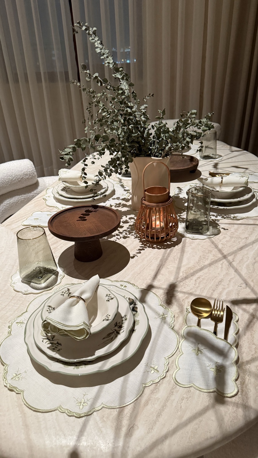 Round dining table set with embroidered scalloped placemats, sage-floral dinnerware and eucalyptus, in front of sheer evening curtains